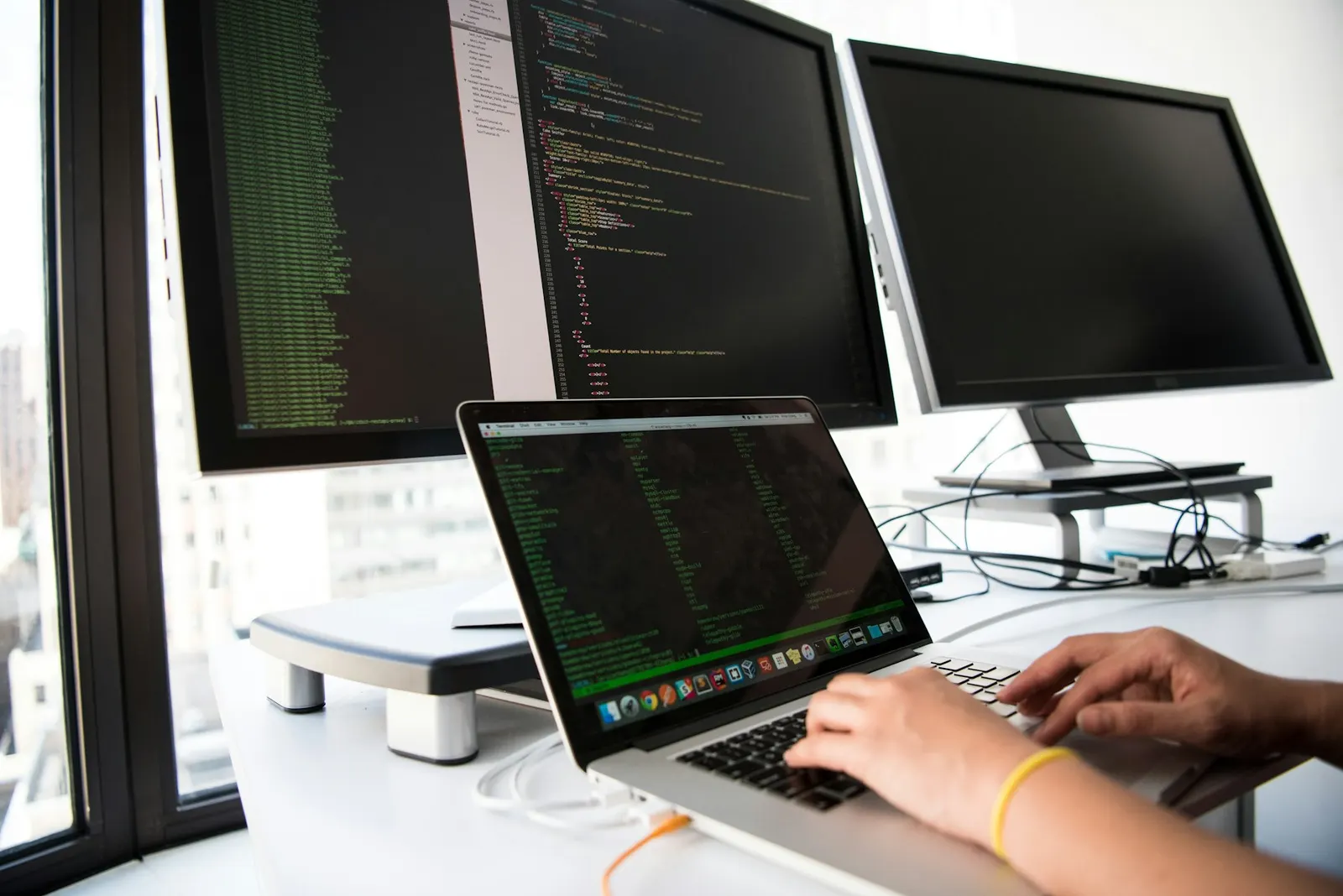 Desarrollador escribiendo código frente a dos monitores y un notebook, con más infraestructura de trabajo